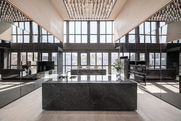 A modern kitchen with a marble island, large windows, and a high ceiling with a unique light fixture.