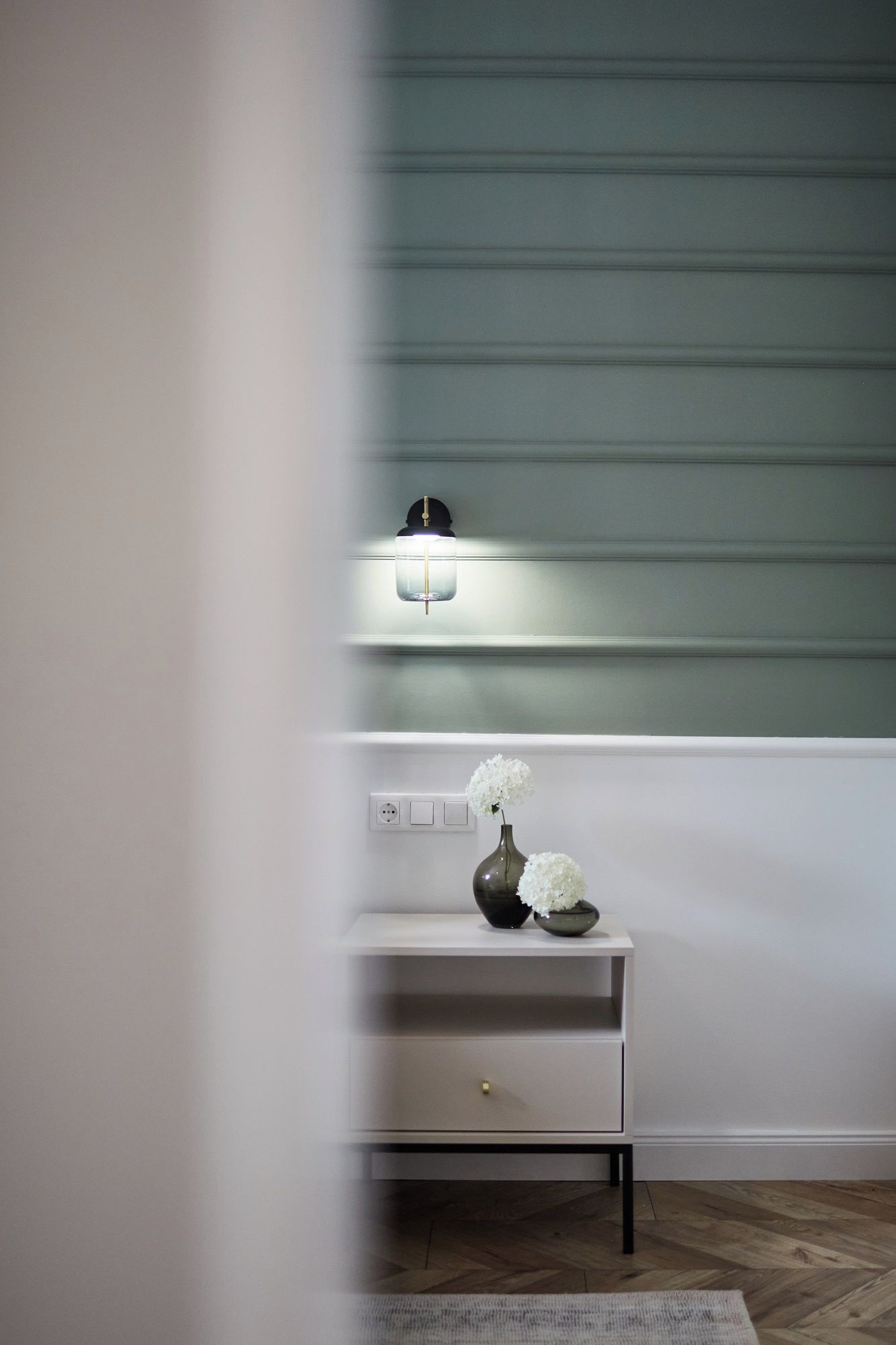 A minimalist room with a white console table, a vase with white flowers, and a wall-mounted light fixture.