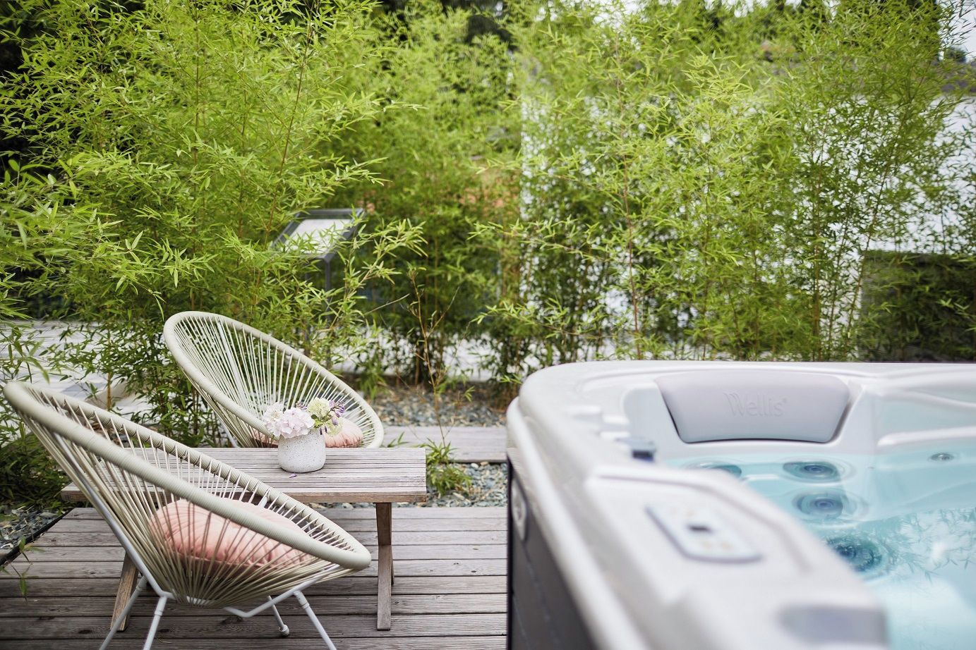 Un bain à remous sur une terrasse en bois, avec deux fauteuils en osier et une petite table sur laquelle est posé un vase de fleurs.