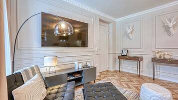 Ein modernes Wohnzimmer mit einem großen Flachbildfernseher, einem schwarzen Sofa, einem runden Hocker und einem Couchtisch aus Holz.