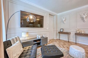 Ein modernes Wohnzimmer mit einem großen Flachbildfernseher, einem schwarzen Sofa, einem runden Hocker und einem Couchtisch aus Holz.