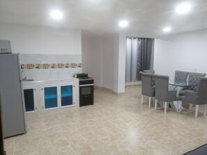 Cocina compacta con refrigerador, estufa y alacenas. Hay un comedor con una mesa de vidrio y cuatro sillas.