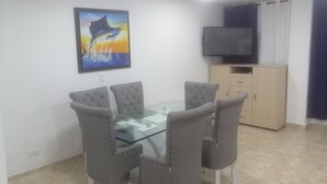 A dining area with a glass table and grey upholstered chairs, a painting on the wall, and a TV on a cabinet. - San Andrés Central Apartment 10 (San Andrés)