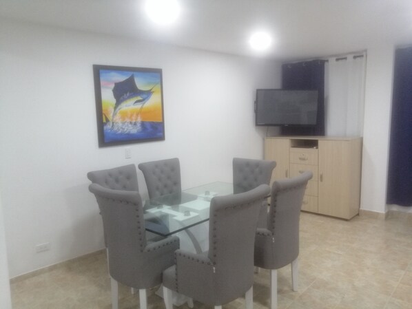 Un comedor con una mesa de vidrio y sillas tapizadas grises, un cuadro en la pared y un televisor sobre un mueble.