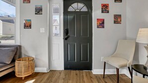 A room with a dark wooden door, a white wall with framed posters, a wicker chair, a sofa, and a lamp. - Nostalgic in Nelsonville: Great back yard for pets (Nelsonville)