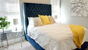 Een slaapkamer met een gestoffeerd hoofdeinde, een bed met wit en geel beddengoed, een klein nachtkastje met een lamp, en een potplant.