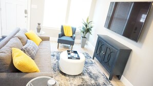 Een woonkamer met een bank, een poef, een fauteuil en een televisie aan de muur.