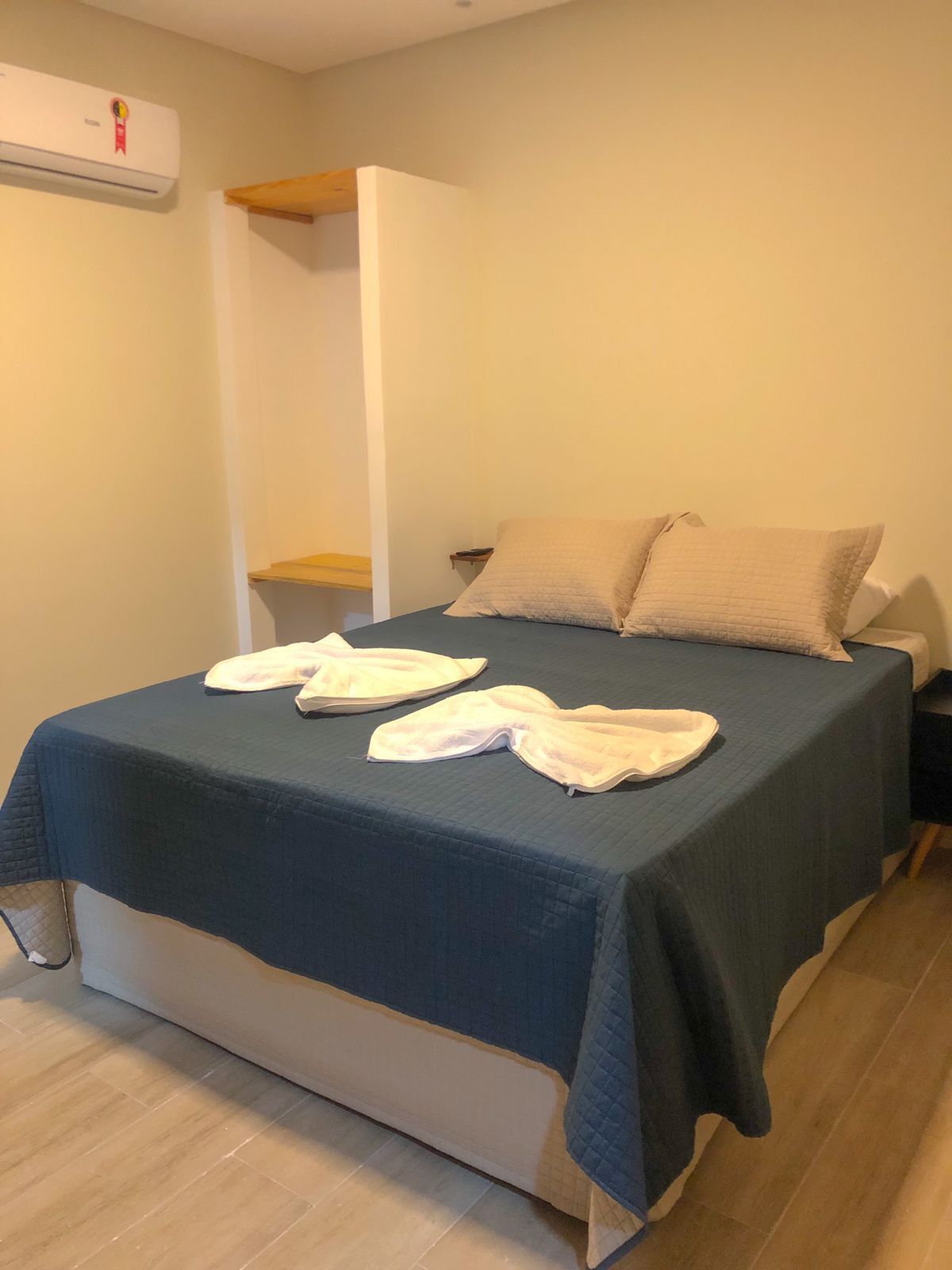 Cama bem arrumada com colcha azul escura e duas almofadas, mesa de cabeceira de madeira com abajur e ar-condicionado fixado na parede.