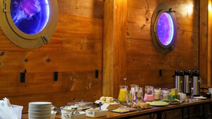 A breakfast buffet with various food items and beverages, wooden walls, and two illuminated circular windows. - Sete Mares (Maragogi)