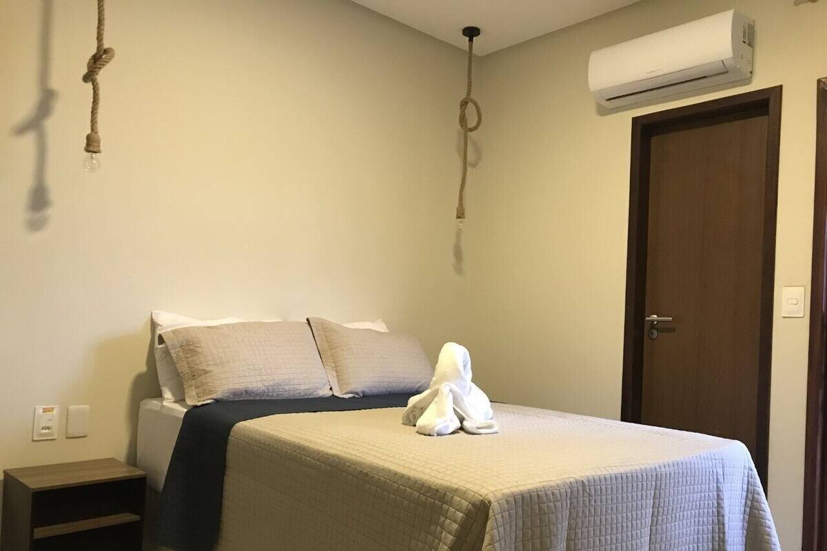 Un lit bien fait, recouvert d’une couverture blanche et surmonté d’une serviette blanche. Une table de chevet en bois se trouve à côté du lit, avec une lampe dessus. On peut voir un climatiseur fixé au mur.