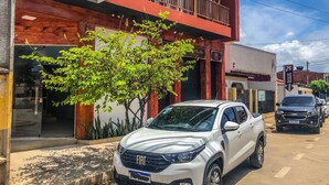 A white FIAT SUV parked on a street with a building featuring a red and white facade, a balcony, and a tree in front. - Sete Mares (Maragogi)