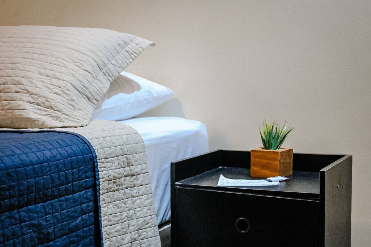 Un lit bien fait, recouvert d’une couette bleue, avec un oreiller beige et une petite table de chevet en bois sur laquelle est posée une plante verte.