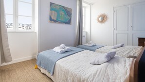 A bedroom with two beds, a window with curtains, a wall painting, and a sunburst mirror. - MA RÉ HOTES        . (Le Bois-Plage-en-Ré)