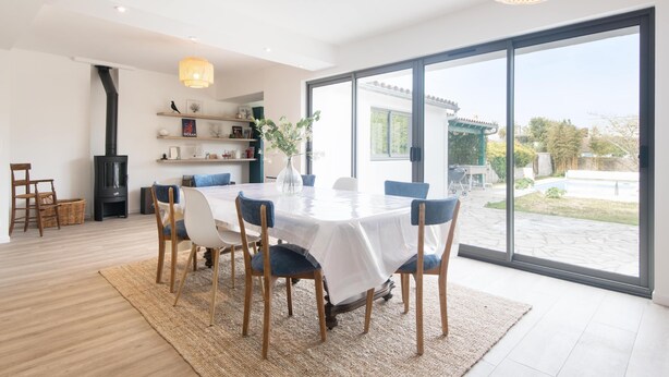 A modern dining area with a round table, blue chairs, and a large window offering a view of a garden and outdoor seating. - MA RÉ HOTES        . (Le Bois-Plage-en-Ré)