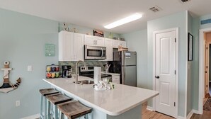 Una cocina con armarios blancos, un refrigerador de acero inoxidable, un microondas y un fregadero. Hay taburetes de madera en la isla de la cocina.