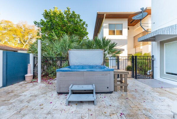 A hot tub with a wooden deck and a small step leading up to it, surrounded by lush greenery and a modern house in the background.