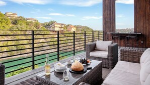Ein Balkon mit Korbmöbeln, einer Weinflasche und einem Weinglas, mit Blick auf einen Fluss und Häuser.
