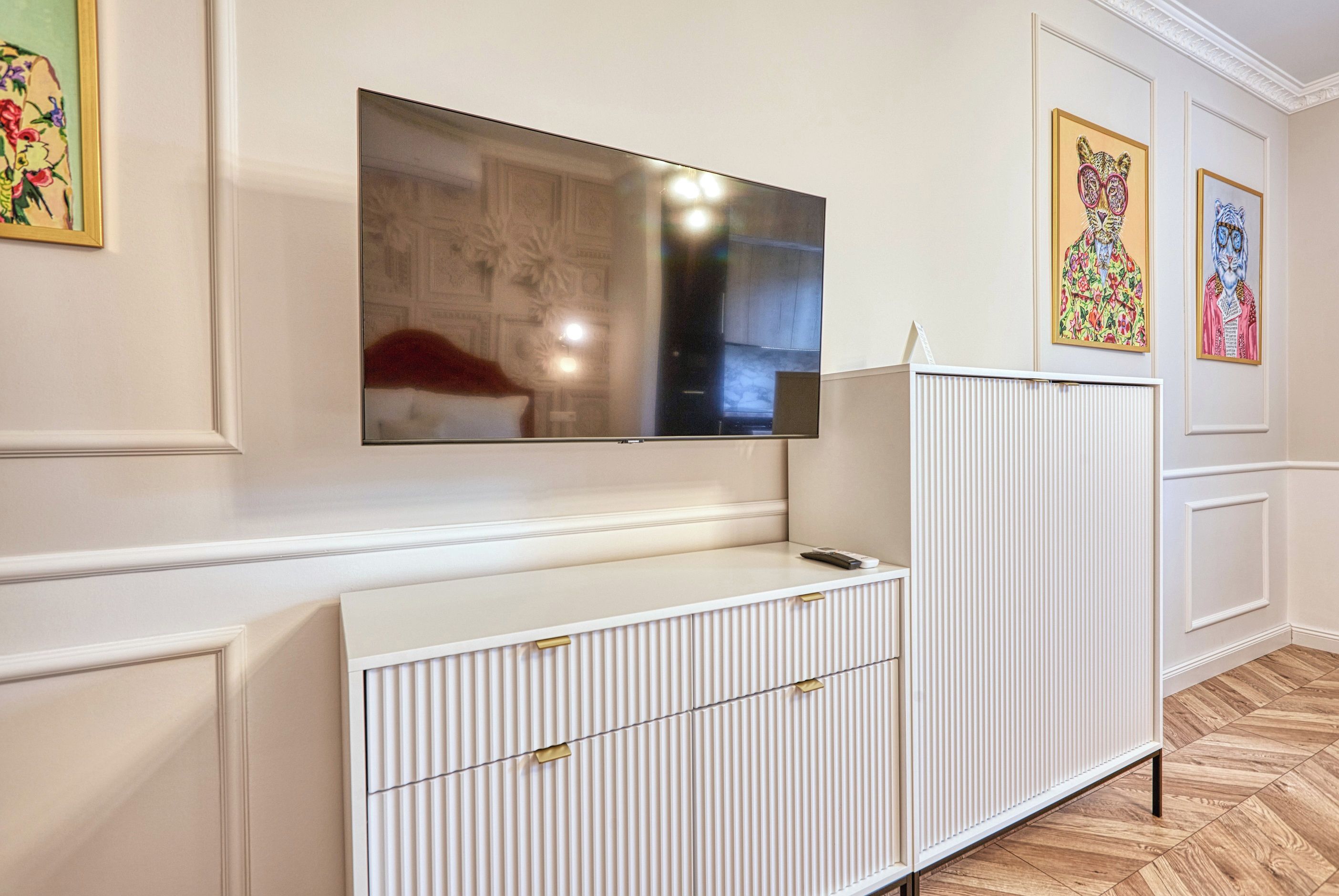 A modern living room with a flat-screen TV mounted on the wall, a white cabinet with drawers, and decorative wall art.