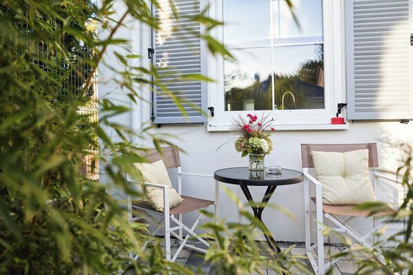 An outdoor seating area with a table and chairs, a window with shutters, and a vase with flowers.