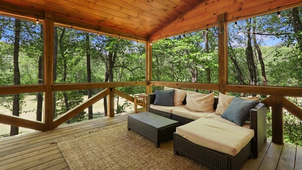 A covered wooden deck with wicker furniture and a view of a forest. - CreekFront Cabin- 4 Acres- FirePit & Place- Game Tables- Private Yet Close to BR (Mineral Bluff)