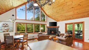 A spacious living room with a fireplace, large windows, and a high ceiling. - CreekFront Cabin- 4 Acres- FirePit & Place- Game Tables- Private Yet Close to BR (Mineral Bluff)