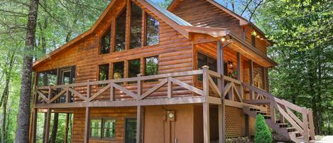 A wooden cabin with a balcony, large windows, and a staircase leading to the upper level.