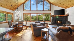 A cozy cabin interior with a fireplace, large windows, wooden floors, and a dining area. - CreekFront Cabin- 4 Acres- FirePit & Place- Game Tables- Private Yet Close to BR (Mineral Bluff)