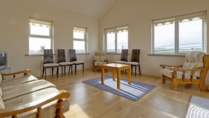 Een ruime woonkamer met een hoge plafond, grote ramen en uitzicht op zee. Er zijn verschillende stoelen en een bank, een houten salontafel en een kroonluchter.