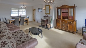 Een woonkamer met een bank, een salontafel, een open haard, een houten kast met boeken, en een eethoek met een tafel en stoelen.