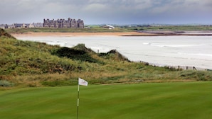 Een golfbaan met een vlaggenstok, een zandstrand en een groot gebouw op de achtergrond.