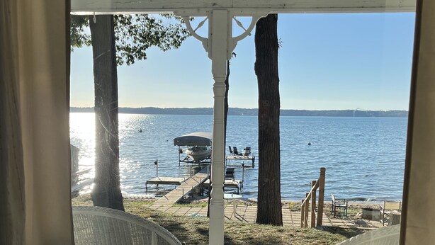 A view from a window overlooking a lakeside area with a dock, chairs, and a boat. - Original Torch Lake Cottage from 1910 (Alden)