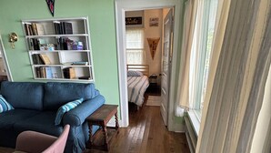 A cozy living room with a blue sofa, a wooden coffee table, and a bookshelf. A door leads to a bedroom with a bed and a window. - Original Torch Lake Cottage from 1910 (Alden)