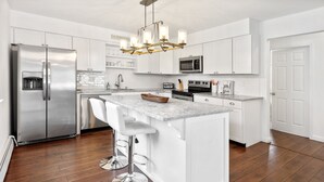 Una cocina moderna con muebles blancos, electrodomésticos de acero inoxidable, una isla de mármol con taburetes altos y una lámpara de araña.