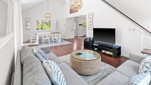 Amplia sala de estar con un televisor grande, un comedor con una mesa y sillas blancas, y una escalera que conduce a un nivel superior.