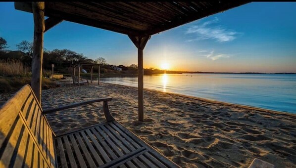 Relax on a serene beach with a shaded bench under a beach hut, surrounded by calm waters and a tranquil atmosphere.