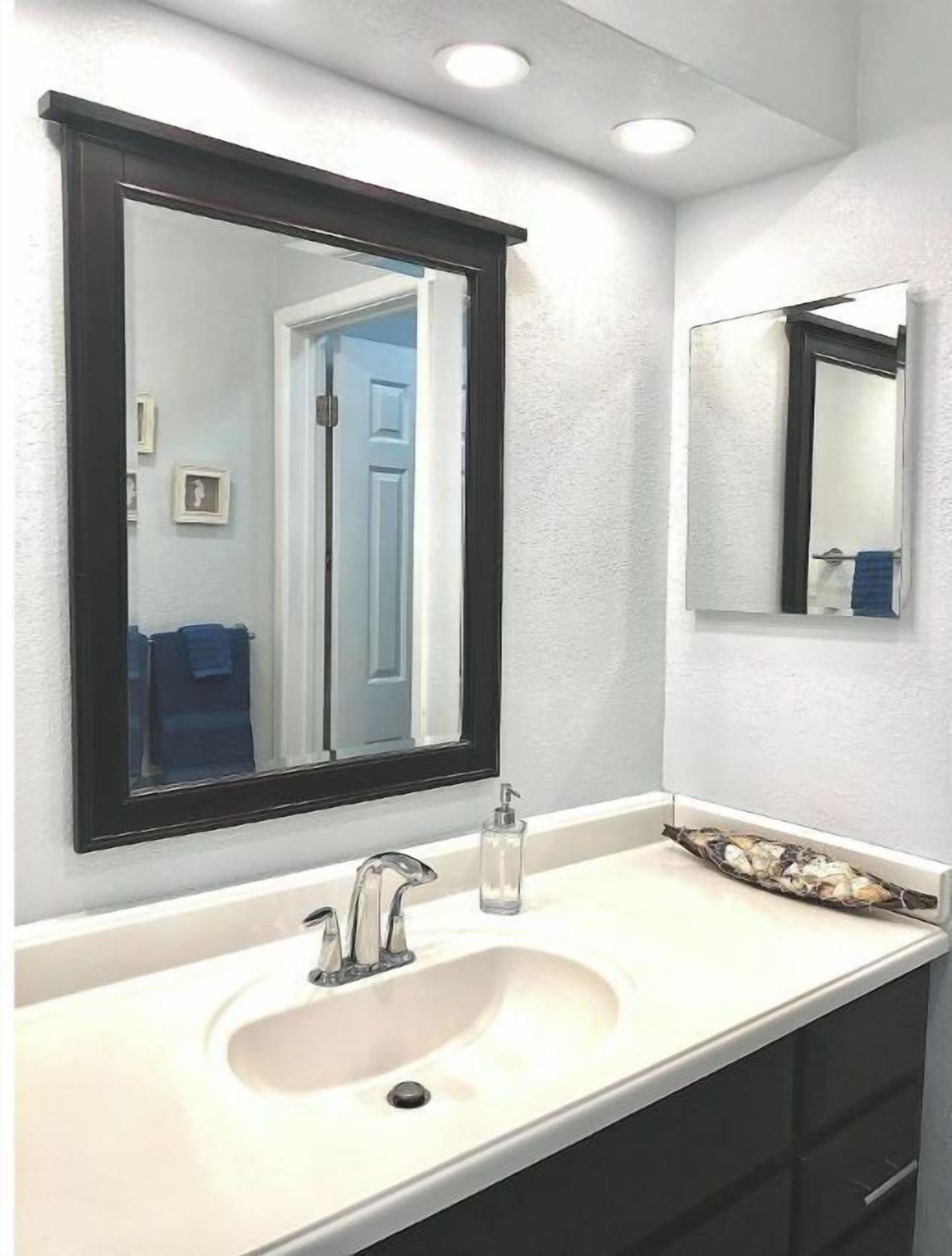 A bathroom with a white sink, a large mirror with a black frame, and a small window.