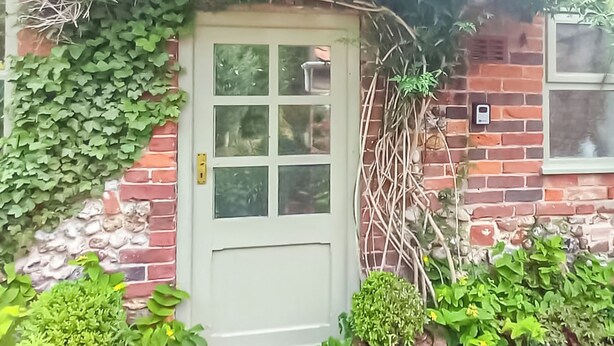Une maison en briques avec une porte blanche, entourée d’une végétation luxuriante et de plantes en pot.