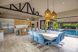 A modern kitchen with a built-in refrigerator, a dining area with a large table and blue chairs, and a pool area with outdoor furniture.