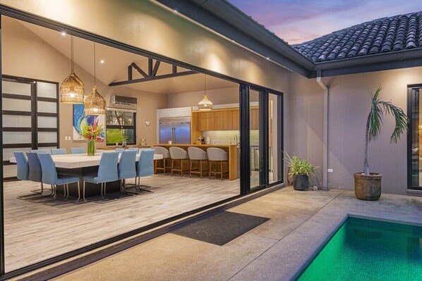 A modern outdoor dining area with a pool, a kitchen visible through glass doors, and a dining table with chairs.