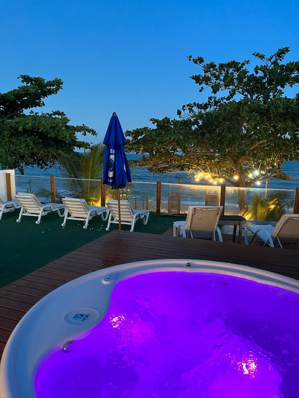 A hot tub with a view of the ocean, surrounded by lounge chairs and an umbrella.