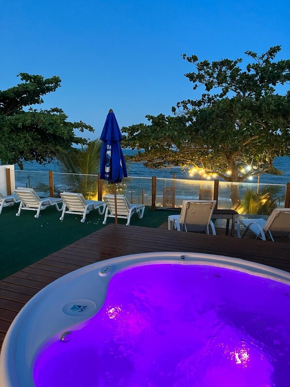 A hot tub with a view of the ocean, surrounded by lounge chairs and an umbrella.