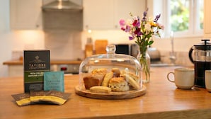 Una encimera de cocina con una caja de café molido Taylor's, una bolsita de té, una campana de cristal sobre pasteles, un florero con flores y una cafetera.