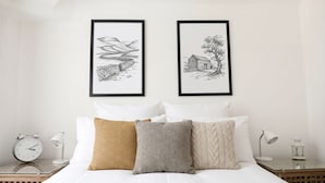Una cama bien hecha con ropa de cama blanca y tres almohadas, una mesita de noche con una lámpara y un reloj, y dos dibujos en blanco y negro enmarcados en la pared.