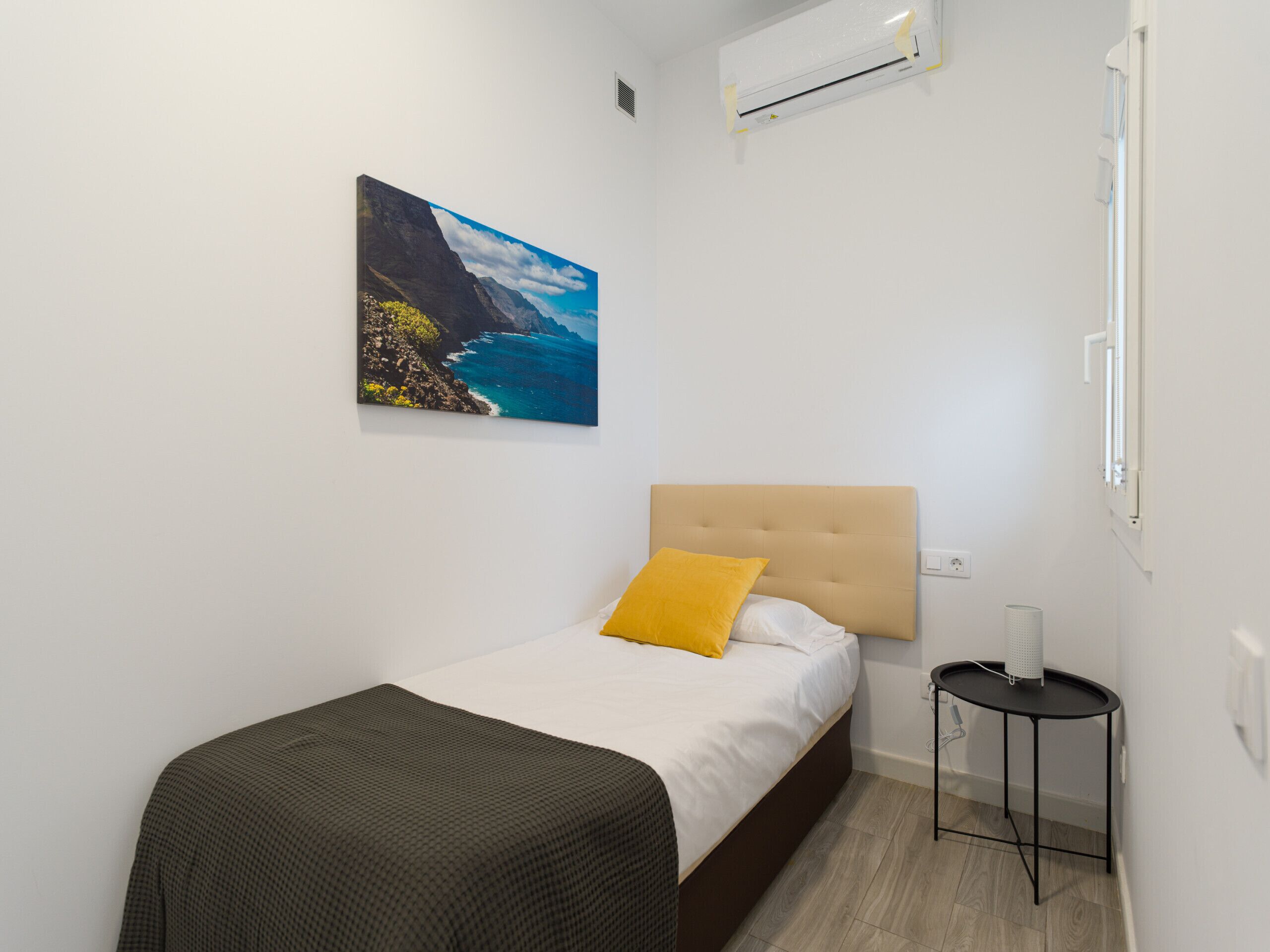 Un dormitorio con cama, una mesita de noche pequeña, un aire acondicionado instalado en la pared y un cuadro de un paisaje costero.