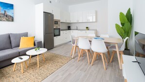 A modern apartment with a grey sofa, a dining table with white chairs, a kitchen with white cabinets, and a large green plant. - Volcano La Colorada By CanariasGetaway (Las Palmas de Gran Canaria)
