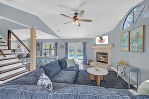 Een moderne woonkamer met een grijze bank, een open haard, een plafondventilator en een eethoek met een glazen tafel.