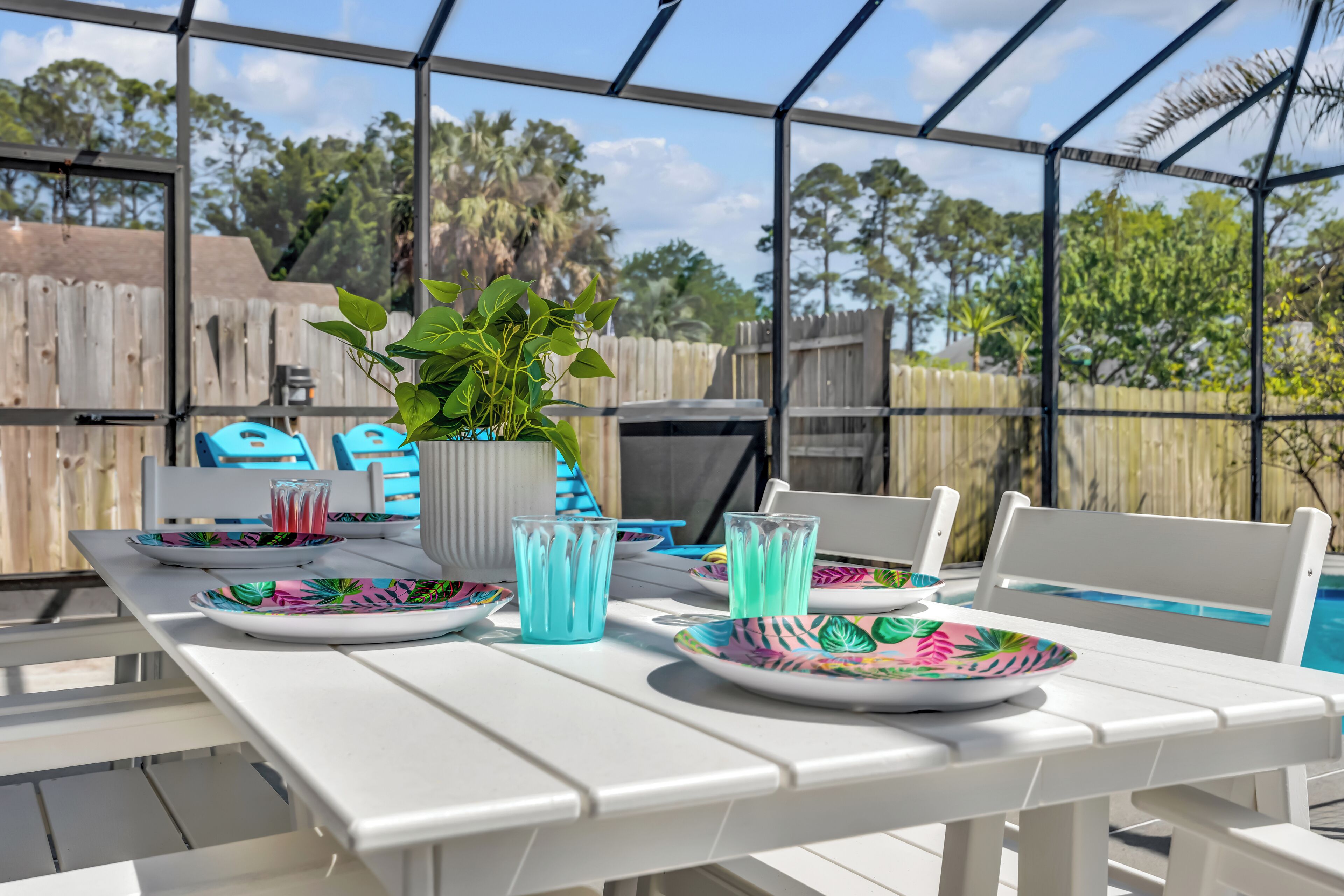 A covered outdoor dining area with a table set for two, a potted plant, and a view of a swimming pool and trees.