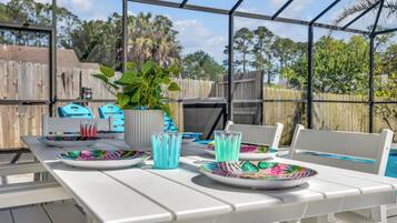A covered outdoor dining area with a table set for two, a potted plant, and a view of a swimming pool and trees.