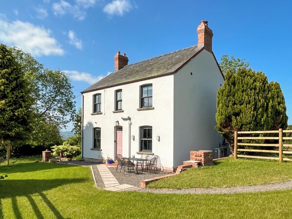 Une maison à deux étages, avec une façade blanche, une cheminée en briques et un petit patio aménagé avec du mobilier d’extérieur.
