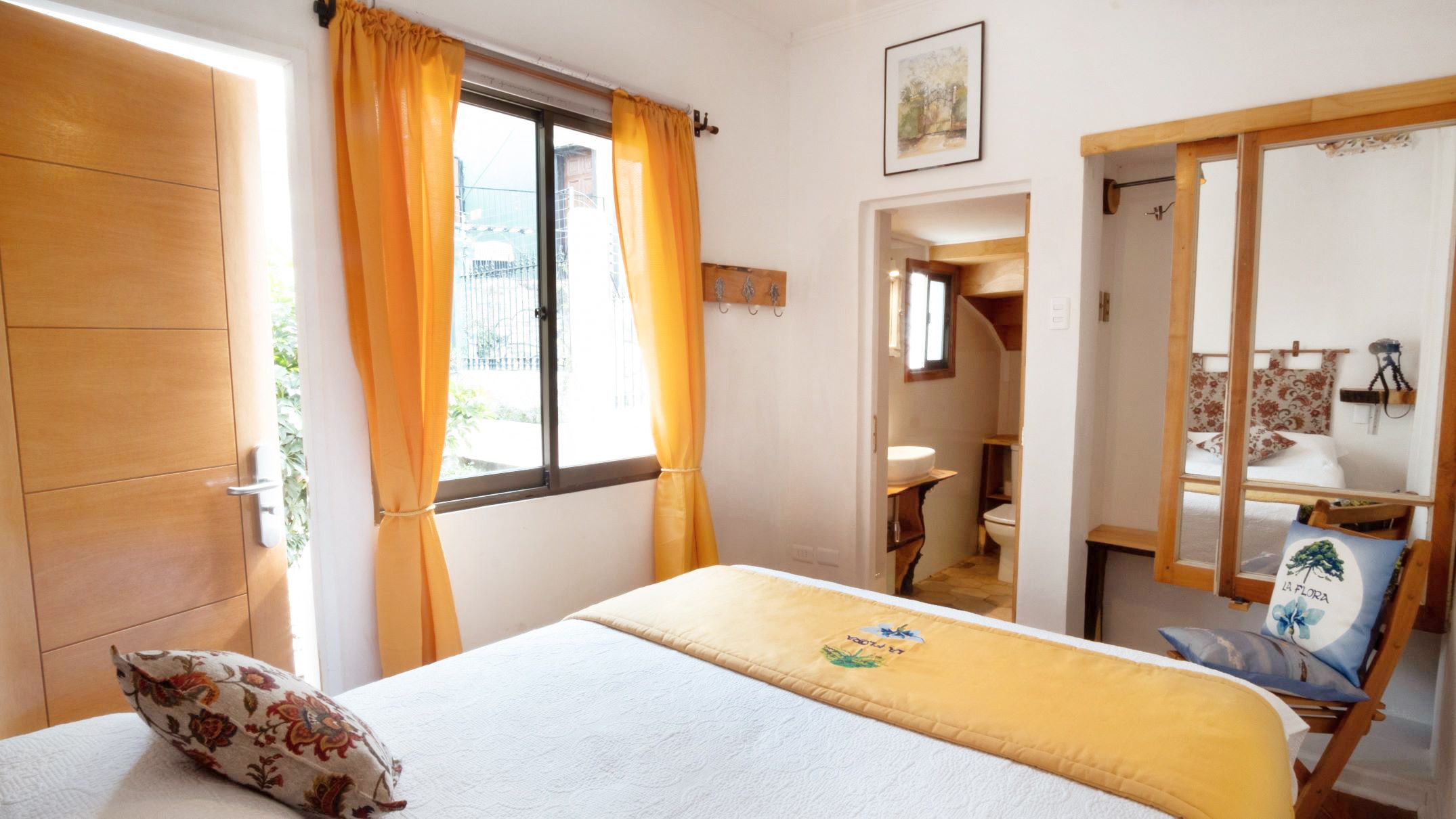 A bedroom with a bed, a window with curtains, a bathroom visible through an open door, and a small seating area with a chair and cushions.
